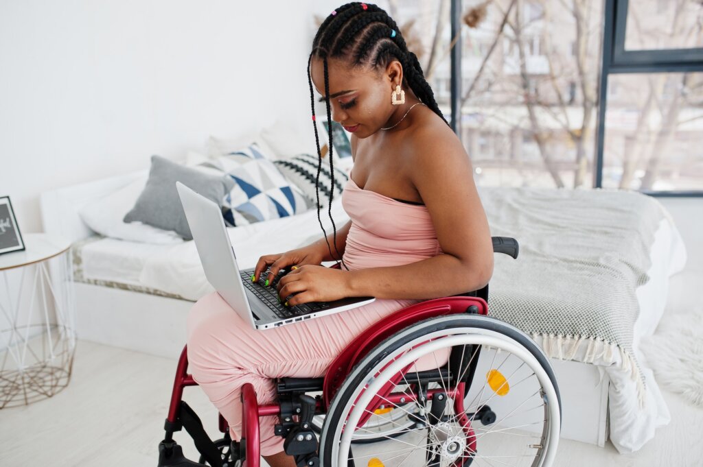 Young disabled African American woman in wheelchair at home working with laptop.